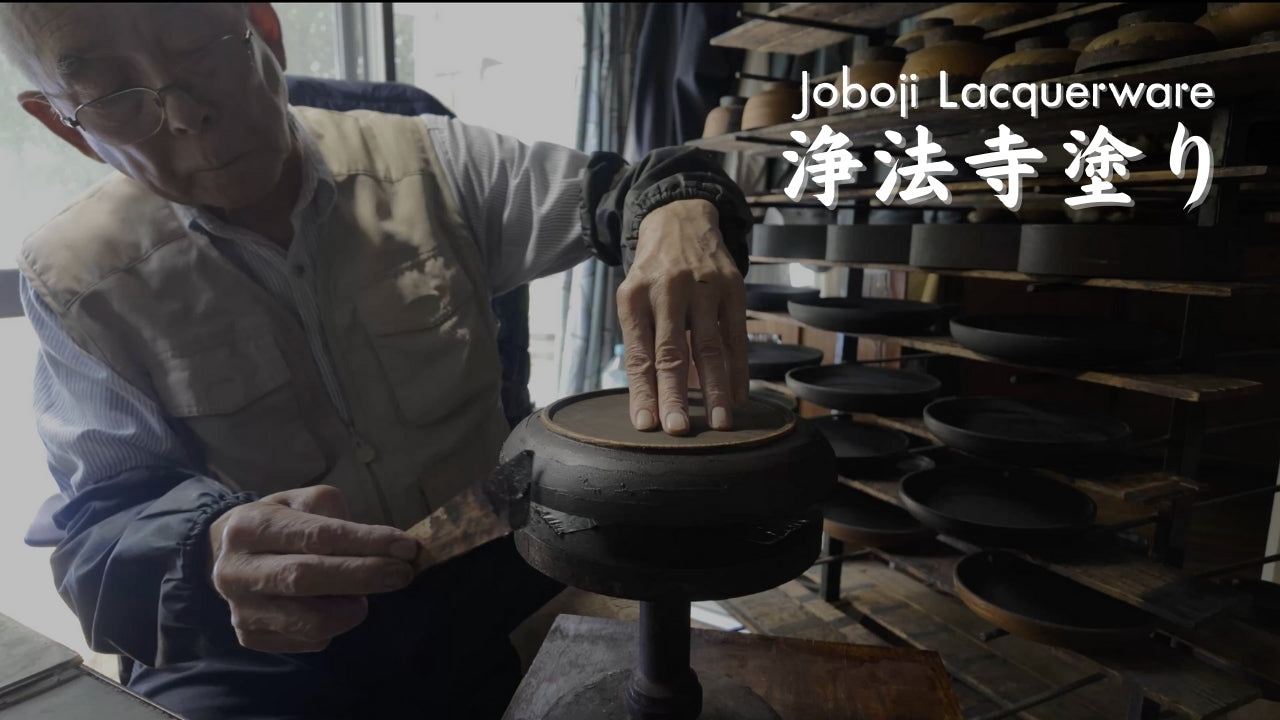 An elderly craftsman lacquers a wooden dish on a turntable, drying racks behind, titled Joboji Lacquerware.
