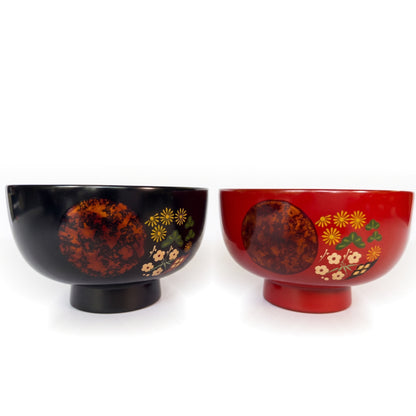 Black and red soup bowls side by side seen slightly from above, each with a copper-mottled circle beside seasonal flowers.