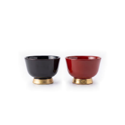 A black lacquer cup and a red lacquer cup stand side by side on gold foot bands, both flared and shown mostly from the side.