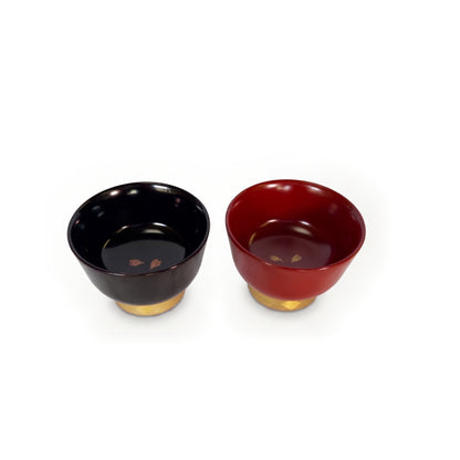 Black and red cups side by side at a high angle, each interior with two cherry blossom petals, bronze on black, gold on red.