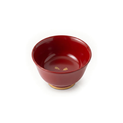 A single red lacquer cup from above, its glossy red interior holding two gold cherry blossom petals near the base's centre.